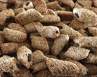 May include: A close-up shot of a pile of sesame cookies. The cookies are oblong, with a light brown exterior completely covered in sesame seeds. A dark filling is visible in some of the cookies, suggesting a date or fruit filling. The cookies are a golden brown color.