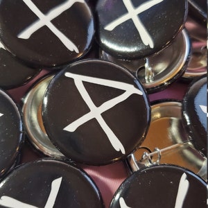 May include: A collection of black button pins with a white, stylized X design. The pins have a glossy finish and metal pin backs. The image shows a close-up view of the pins, with some overlapping.
