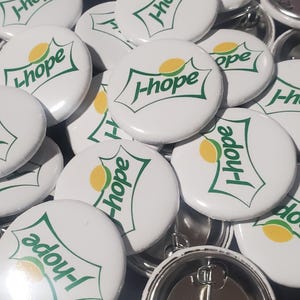 May include: A collection of round white pin-back buttons featuring a green graphic logo with the text J-hope and a yellow oval shape. Each button has a smooth, glossy finish and a metal pin mechanism visible on the back of the bottom-most piece.