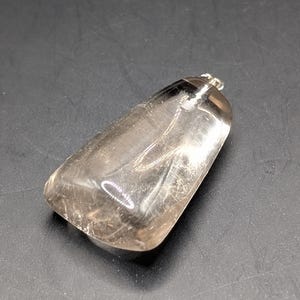 Peut inclure: Un pendentif rectangulaire transparent avec une bélière argentée. Le pendentif est de couleur fumée translucide avec des inclusions internes. La forme est arrondie en haut et se rétrécit légèrement vers le bas. Le pendentif est probablement en cristal ou en pierre précieuse.