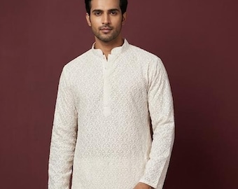 Pode incluir: Um conjunto kurta pijama bordado creme. A kurta de manga comprida apresenta uma gola mandarim e intrincados padrões florais. As calças de pijama a condizer são de cor creme lisa. A roupa é usada por uma pessoa em frente a um fundo bordô.