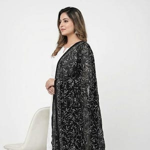 May include: A black sheer dupatta with intricate silver floral embroidery. The dupatta is draped over a white outfit. The design features a detailed pattern of leaves and vines, creating a delicate and elegant look.