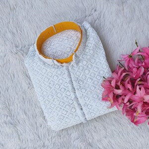 Pode incluir: Top branco de renda com colarinho amarelo e fecho de bot&otilde;es. O top est&aacute; dobrado e colocado sobre uma superf&iacute;cie branca e felpuda.