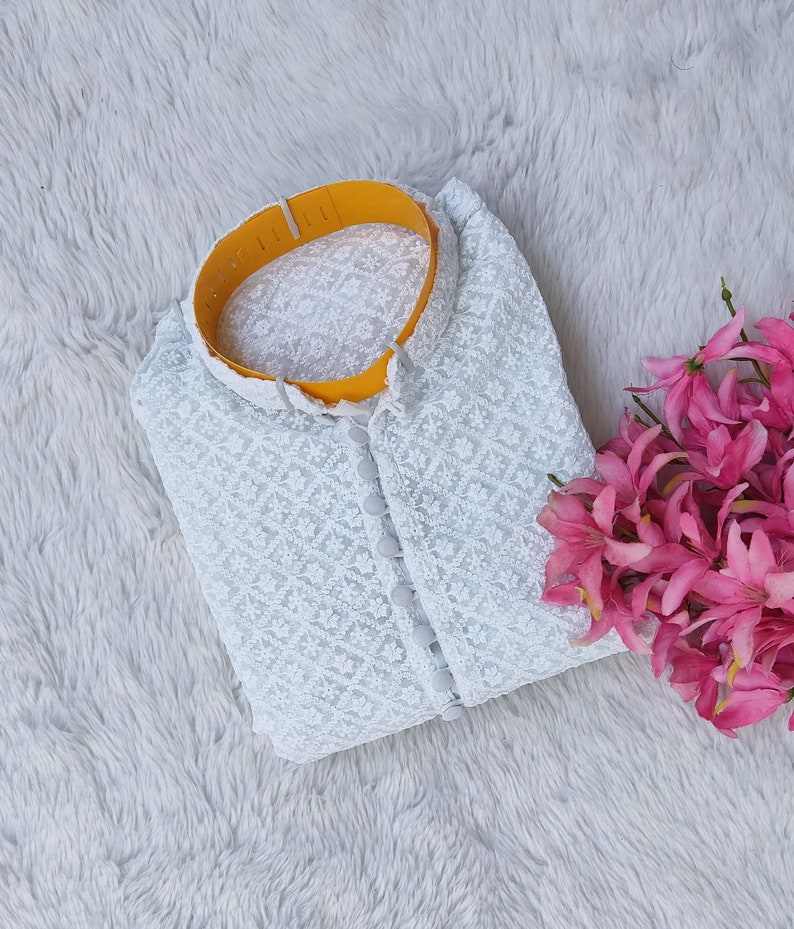 Pode incluir: Top branco de renda com colarinho amarelo e fecho de bot&otilde;es. O top est&aacute; dobrado e colocado sobre uma superf&iacute;cie branca e felpuda.