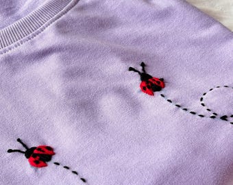 Puede incluir: Una camiseta de color lavanda con tres mariquitas bordadas. Cada mariquita tiene patas y antenas negras, un cuerpo rojo con manchas negras y un contorno negro. Las mariquitas están bordadas con una línea discontinua negra que se arrastra detrás de ellas.