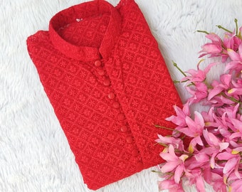 Pode incluir: Uma blusa vermelha bordada com botões e gola mandarim. A blusa está dobrada e colocada sobre uma superfície branca e fofa.