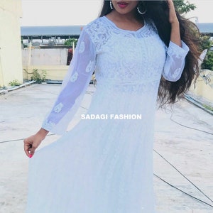 May include: A white sheer long-sleeved dress with intricate embroidery. The dress has a round neckline and a flowing skirt. The text "SADAGI FASHION" is visible on the dress.