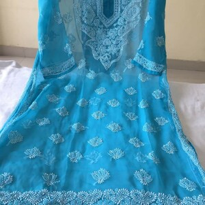 May include: A turquoise blue sheer tunic with white embroidery. The embroidery features floral and paisley designs. The tunic has a round neckline and three-quarter sleeves.