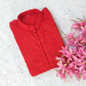 Pode incluir: Uma blusa vermelha bordada com bot&otilde;es e gola mandarim. A blusa est&aacute; dobrada e colocada sobre uma superf&iacute;cie branca e fofa.