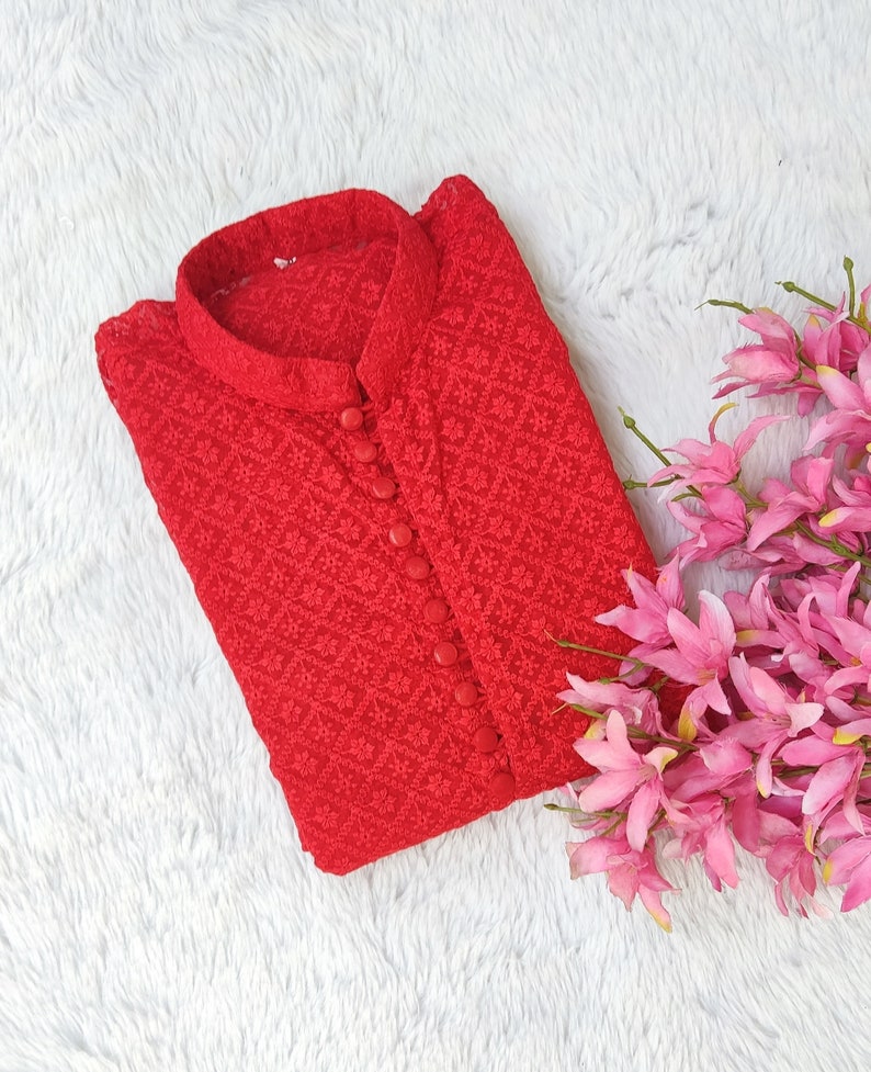 Pode incluir: Uma blusa vermelha bordada com bot&otilde;es e gola mandarim. A blusa est&aacute; dobrada e colocada sobre uma superf&iacute;cie branca e fofa.
