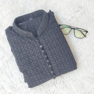 Pu&ograve; includere: Una kurta nera con motivo, colletto alla coreana e chiusura con bottoni. La kurta &egrave; piegata e appoggiata su una superficie bianca e pelosa.