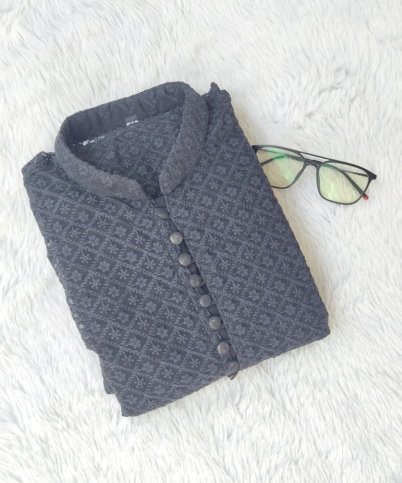 Pu&ograve; includere: Una kurta nera con motivo, colletto alla coreana e chiusura con bottoni. La kurta &egrave; piegata e appoggiata su una superficie bianca e pelosa.