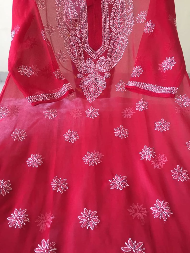 May include: A red sheer tunic with white embroidery. The embroidery features a floral pattern and a paisley design. The tunic has a round neckline and three-quarter sleeves.
