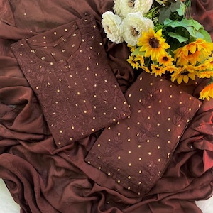 May include: A dark brown fabric set featuring a tunic and matching material with intricate tonal embroidery and small gold-coloured polka dot accents. The ensemble is displayed alongside a bouquet of white and yellow artificial flowers.