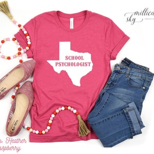May include: A heather raspberry t-shirt with the state of Texas in white, and the words "SCHOOL PSYCHOLOGIST". The shirt is paired with blue jeans and pink flats. A decorative beaded garland with a tassel is also included.