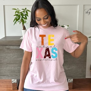 May include: Pink t-shirt with the text "Texas School Psychologist" in colorful, distressed lettering.