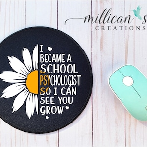 May include: A black round mouse pad with a white daisy and the text "I became a school psychologist so I can see you grow" in white with a yellow center to the daisy.