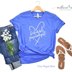 May include: A royal blue t-shirt with a white heart and the text "School Psychologist" inside the heart. The shirt is on a white background with a pair of white sandals and a pair of blue jeans.