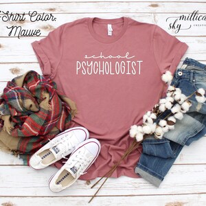 May include: Mauve t-shirt with the text "school psychologist" in white lettering. The shirt is laid flat on a white wooden surface with a pair of white sneakers, a plaid scarf, and a bunch of cotton bolls.