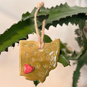 Puede incluir: Adorno de cerámica con forma de Ohio, con un corazón rojo y detalles florales. El adorno es de color amarillo dorado y cuelga de una cuerda de hilo natural. El fondo presenta hojas verdes, creando un contraste natural.