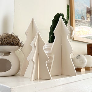 May include: White ceramic Christmas tree decorations on a white mantel with a white ceramic figurine and a brown bird's nest.