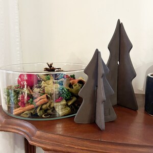 Puede incluir: Dos adornos de árbol de Navidad de cerámica marrón sobre una mesa de madera. Un cuenco de vidrio lleno de popurrí se encuentra delante de los adornos.