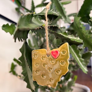 Puede incluir: Adorno de cerámica con forma de Ohio, con un corazón rojo y diseños florales. El adorno es de color amarillo dorado, suspendido por una cuerda de hilo natural. El fondo muestra una planta verde.