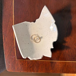 May include: A speckled, off-white ceramic ring dish in a leaf shape, holding two gold rings. The dish sits on a dark wooden surface. The rings are a simple band and a band with small stones.