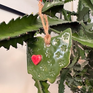 May include: A green ceramic ornament in the shape of the state of Ohio, with a red heart and floral patterns. The ornament is suspended by a natural twine string and is surrounded by green plant foliage.