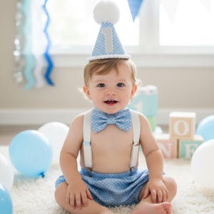 May include: A baby wearing a light blue polka dot outfit, including a bow tie, suspenders, and shorts. A matching party hat with a white pom-pom and the number "1" is on the baby's head. Balloons in shades of blue and white are in the background.