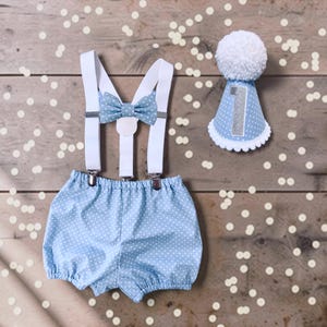 May include: A light blue bloomer set with white polka dots, featuring white elastic braces and a matching bow tie. Beside the bloomers is a matching light blue party hat with white polka dots, a white pom-pom top, and a silver glitter number 1.