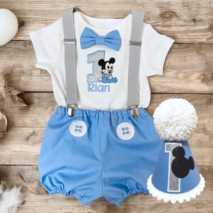 May include: A white short-sleeved bodysuit with a blue bow tie, a glittery number one, a Mickey Mouse graphic, and the name Rian, paired with light blue shorts featuring two white buttons and grey braces, plus a matching blue party hat.