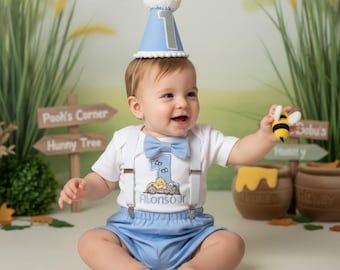 May include: A baby in a light blue outfit with braces, a bow tie, and a matching party hat with a silver number 1. The outfit features a Winnie the Pooh graphic and the name "Alonso Jr." The baby holds a toy bee.