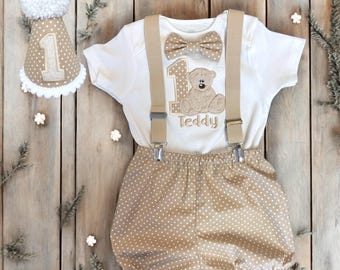 May include: A beige and white first birthday outfit set. The set includes a white onesie with a teddy bear applique, suspenders, a bow tie, and polka dot shorts. A matching party hat with a pom-pom and the number "1" completes the ensemble.