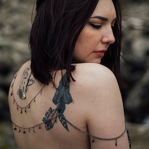 Peut inclure: Une femme aux cheveux courts et fonc&eacute;s et un piercing au nez porte un collier d&eacute;licat en argent avec des breloques en forme d'&eacute;toile et de croissant de lune. Le collier est superpos&eacute; &agrave; un tatouage sur son dos qui repr&eacute;sente un oiseau, une plume et une feuille.