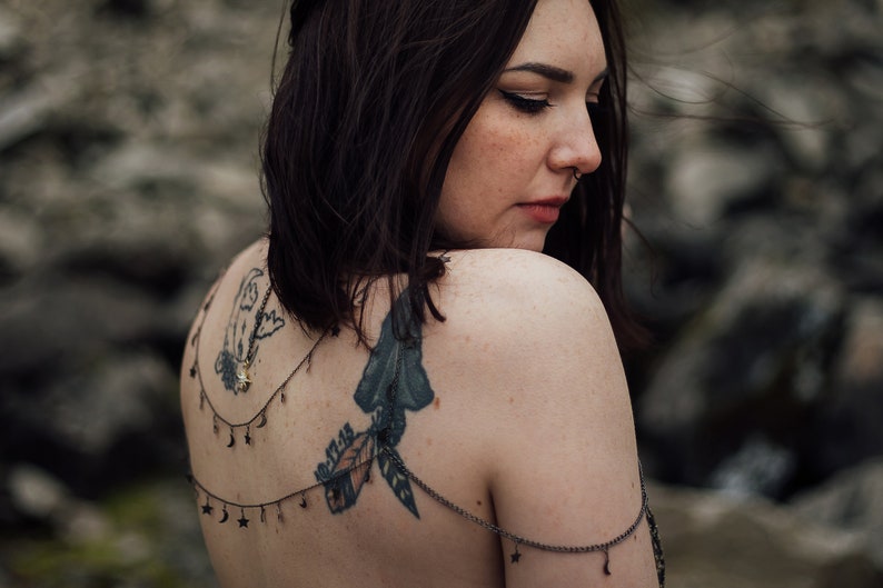 Peut inclure: Une femme aux cheveux courts et fonc&eacute;s et un piercing au nez porte un collier d&eacute;licat en argent avec des breloques en forme d'&eacute;toile et de croissant de lune. Le collier est superpos&eacute; &agrave; un tatouage sur son dos qui repr&eacute;sente un oiseau, une plume et une feuille.