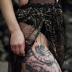 Peut inclure: Une robe noire transparente avec des paillettes dor&eacute;es et un d&eacute;tail de cha&icirc;ne noire. La robe est port&eacute;e par une personne avec un grand tatouage sur la cuisse d'un cr&acirc;ne avec des d&eacute;tails floraux.