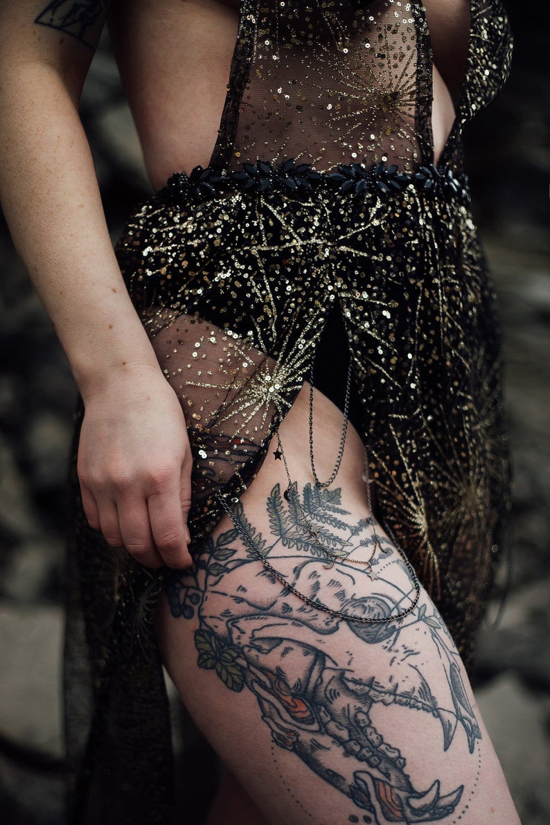 Peut inclure: Une robe noire transparente avec des paillettes dor&eacute;es et un d&eacute;tail de cha&icirc;ne noire. La robe est port&eacute;e par une personne avec un grand tatouage sur la cuisse d'un cr&acirc;ne avec des d&eacute;tails floraux.