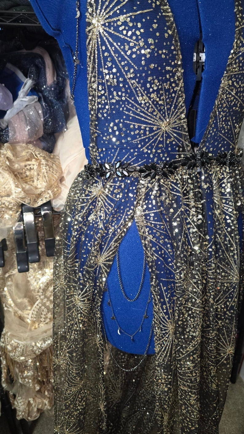 Peut inclure: Une robe en maille noire avec des &eacute;toiles dor&eacute;es scintillantes et une ceinture noire. La robe a un d&eacute;collet&eacute; en V profond et une longue jupe fluide.