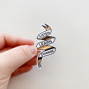 May include: A white sticker with a brown and orange ribbon design. The ribbon is curled and has the words "LISTEN", "LEARN", and "CHANGE" written on it.
