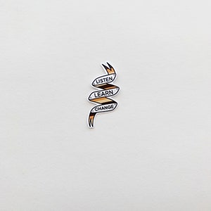 May include: A white sticker with a brown and black ribbon design. The ribbon is curled and has the words "Listen, Learn, Change" printed on it.