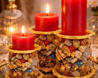 May include: Three lit red pillar candles on decorative holders. The holders are cylindrical, encrusted with colourful stones, and trimmed with gold filigree. The candles are on a gold-rimmed tray.