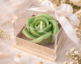 May include: A green rose-shaped candle with gold detailing, presented in a clear acrylic box with a gold base. The candle is tied with a white satin bow. Decorative floral elements and gold heart-shaped confetti are scattered around.