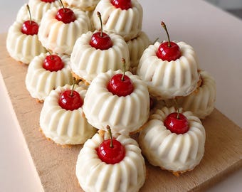 May include: A wooden board displays a stack of mini bundt cakes, each crowned with a vibrant red cherry. The cakes feature a white, ridged design, and the cherries have green stalks. The cakes are arranged on a light brown wooden board.