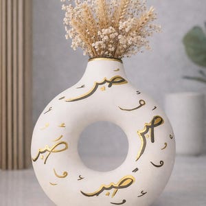 Puede incluir: Jarrón de cerámica blanca con un diseño único en forma de donut, adornado con caligrafía árabe dorada y negra. El jarrón contiene trigo seco y pequeñas flores blancas, creando una estética minimalista y elegante.