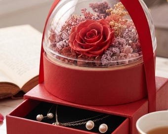 May include: A red jewellery box with a drawer, topped with a glass dome containing a red rose and dried flowers. A red ribbon forms a handle. The drawer contains a necklace and earrings with pearls.