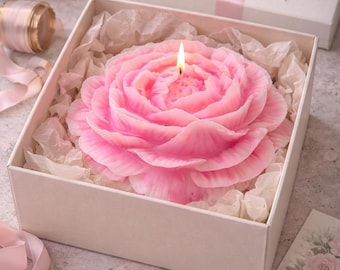 May include: A pink, rose-shaped candle with a lit wick is presented in a white box with tissue paper. A gift box with a pink ribbon and a tag reading "For You" are also visible. The candle is surrounded by rose petals and other floral elements.