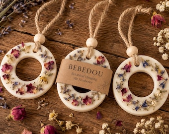 May include: Three cream-coloured, circular air fresheners with dried flowers and a jute string for hanging. One has a small brown tag that reads "BEBEDOU Botanical Hanging Air Freshener". The air fresheners are on a wooden surface with dried flowers.