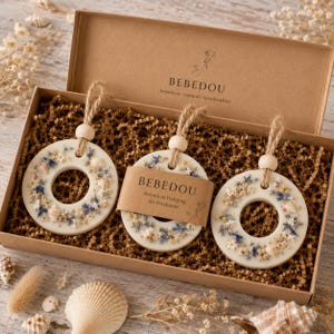 May include: Three round, cream-colored air fresheners with a central hole, adorned with dried flowers and seashells, are displayed in a cardboard box. Each has a wooden bead and twine for hanging. The box is filled with brown crinkle paper, and the brand name "BEBEDOU" is visible.