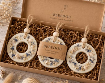 May include: A set of three cream-coloured, circular air fresheners with a central hole, decorated with dried flowers and seashells, presented in a cardboard box. Each has a wooden bead and twine for hanging. The box contains brown crinkle paper, and the brand name "BEBEDOU" is visible.